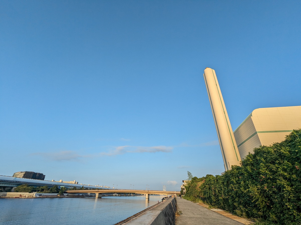 川沿いの防波堤のある道。道沿いに大きな煙突のある建物が立っている。空は真っ青。